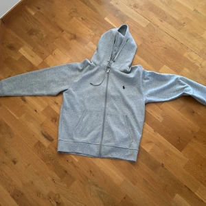 Grå hoodie från Ralph Lauren - Snygg grå hoodie från Ralph Lauren med dragkedja och klassisk logga på bröstet. Tröjan har huva med snörning, två fickor framtill och är tillverkad i mjuk bomullsmix. Perfekt för chill dagar eller när du vill ha en clean streetstyle.