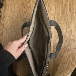 Svart datorväska från Don Donna - Stilren svart datorväska från Don Donna med två handtag och dragkedja. Väskan är tillverkad i skinn och har ett rymligt fack med ljus insida och flera innerfickor för smart organisering. Perfekt för att bära laptop och viktiga prylar till plugget eller jobbet.