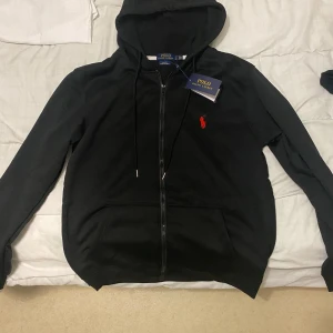 Svart hoodie från Polo Ralph Lauren - Svart hoodie från Polo Ralph Lauren med dragkedja, huva och snörning. Klassisk röd logga broderad på bröstet och stora fickor framtill. Perfekt för en chill och stilren look. Helt ny o använd säljer för storlek passade ej.