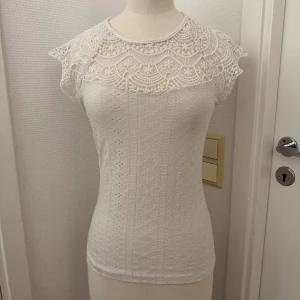 Vit spetstopp XS stretch - Supersöt vit topp med broderad spets upptill och små hålmönster över hela tyget. Toppen har korta ärmar, rund halsringning och knapp i nacken. XS stretch i tyget. Kan hämtas i Hägersten. 