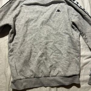 Grå sweatshirt från Kappa - Grå sweatshirt från Kappa med klassisk logga på bröstet och svarta detaljer längs ärmarna. Tröjan har rund halsringning, ribbade muddar och är tillverkad i mjuk bomullsblandning. Perfekt för en sportig och avslappnad stil.