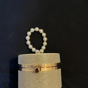 Glittrig kuvertväska med pärlhandtag - Superfin rund kuvertväska i guld med glittriga detaljer och ett handtag av stora pärlor. Väskan har hårt skal och stängs med ett snyggt guldfärgat spänne. Perfekt accessoar för att sticka ut och ge din outfit extra bling. 
