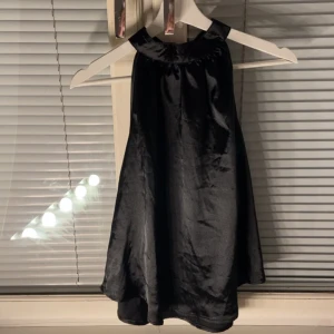 Svart halterneck topp i satin - Snygg svart halterneck topp i glansigt satinmaterial. Toppen har en hög halsringning och är ärmlös med en lös passform som faller fint. Perfekt för dig som vill ha en stilren och trendig look.