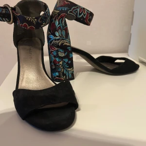 Blommiga klackskor från Tamaris - Snygga klackskor från Tamaris med svart mocka framtill och bred klack täckt av färgglatt blommigt mönster i blått, rött och guld. Skorna har öppen tå, bred rem över foten och justerbart spänne runt ankeln. Perfekta för dig som vill sticka ut med coola detaljer.
