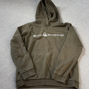 Sail Racing - olivfärgad hoodie (junior)  med tryck - Olivgrön hoodie från Sail Racing (junior) med vit logga framtill och liten logga på huvan. Klassisk känguruficka, ribbade muddar och mjuk insida. Perfekt för dig som gillar en avslappnad och sportig stil.