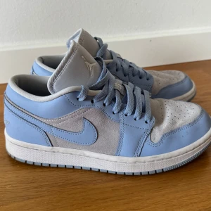 Nike Air Jordan 1 Low blå/grå sneakers - Säljer ett par Nike Air Jordan 1 Low i en snygg ljusblå och grå färgkombination. Skorna har ovandel i mocka och skinn, klassisk snörning och broderad Air Jordan-logga på hälen. Perfekta för dig som vill sticka ut med stilrena sneakers.