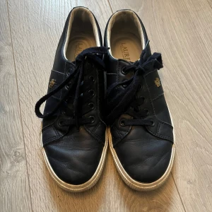 Marinblåa sneakers från Ralph Lauren - Marinblåa sneakers från Ralph Lauren med klassisk design. Skorna har snörning, rund tå och vit sula. Tillverkade i skinn med guldfärgad RL-detalj på sidan. Använda fåtal gånger o är i väldigt bra skick, däremot är de smutsiga på sulorna, men inget som är svårt att få bort (räcker med att torka av de med blött papper). Köpta på boozt för något år sedan, nypris ca. 800kr. Storlek: 38,5
