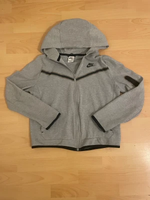 Grå Nike tech zip hoodie - Snygg grå zip hoodie från Nike i storlek 158-170cm. Tröjan har hel dragkedja, huva och svarta detaljer vid muddar och bröstet. Materialet är mjuk bomull och polyester, perfekt för chill eller träning. Nike-logga tryckt på bröstet och praktiska fickor framtill.