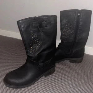 Svarta boots med nitar och spännen - Säljer ett par svarta boots i skinn med coola nitar i guld på skaftet och snygga spänndetaljer. Dragkedja på sidan för enkel påtagning och robust klack. Perfekt för dig som gillar en edgy stil och vill sticka ut.
