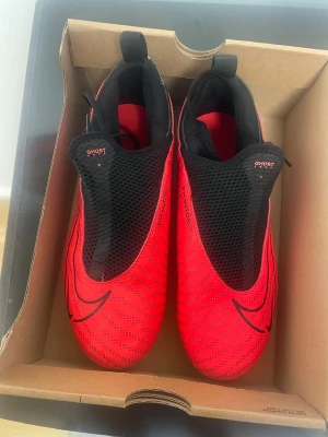 Nike Phantom GT fotbollsskor röd/svart - Nike Phantom GT fotbollsskor i röd och svart med vita detaljer. Skorna har en strömlinjeformad design, syntetiskt material och snörning som döljs under mesh-panelen. Sulan har dobbar för gräsplan och en rund tå. Snygga mönster och Nike-logga på sidan och hälen.