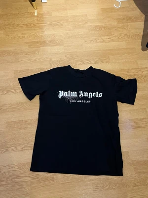 Svart Palm Angels t-shirt - Svart t-shirt från Palm Angels med vit logotyp och Los Angeles-tryck på bröstet. Klassisk rund hals och korta ärmar. Snygg streetwear-stil med en loose passform, perfekt för dig som gillar trendiga märken.