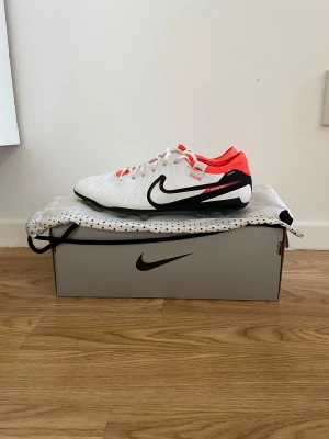 Nike Tiempo Legend 9 Elite FG fotbollsskor - Nike Tiempo Legend 9 Elite FG fotbollsskor i vitt med svarta och röda detaljer. Skorna är i storlek 41. Ovandelen är i skinn och mesh för bästa känsla och passform. Säljes med originalpåse och låda. I väldigt fint skick. Nypris ca 2800 kr.