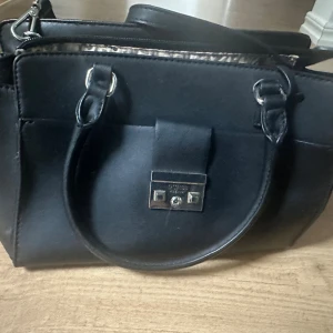 Svart handväska/axelväska från Guess - Snygg svart handväska från Guess med stilren design och silverfärgad metallplatta framtill. Väskan har rundade handtag och insidan är fodrad med leopardmönster. Perfekt storlek för dina essentials och har praktiska fack med dragkedja.