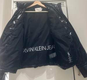 Svart pufferjacka Calvin Klein Jeans - Snygg svart pufferjacka från Calvin Klein Jeans med glansig finish och quiltad design. Jackan har dragkedja framtill, ståkrage och logga både på bröstet och ryggen. Materialet är polyester och jackan är långärmad med normal passform.
