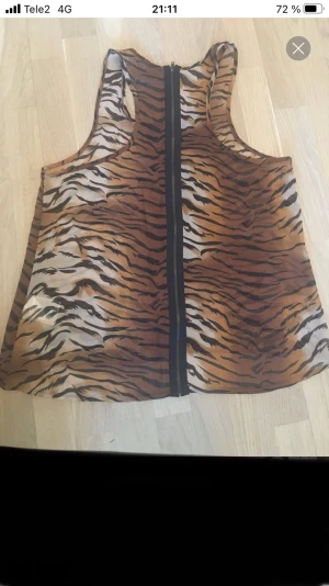 Tigerprintad ärmlös linnetopp Saints & Mortals - Säljer en cool ärmlös linnetopp från Saints & Mortals i transparent syntetmaterial med tigerprint i brunt, beige och svart. Toppen har rund hals och dragkedja längs hela ryggen för extra edge. Perfekt för dig som gillar djurmönster och vill sticka ut.