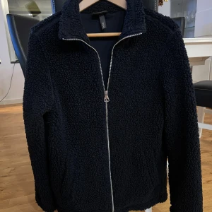 Svart teddyjacka från H&M - Mysig svart teddyjacka från H&M med regular fit. Jackan har en hög krage, hel dragkedja framtill och två sidofickor. Materialet är fluffigt och ger en cool, avslappnad vibe. Perfekt för att hålla värmen under kyliga dagar.