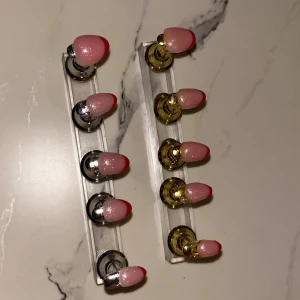 Rosa och röda press-on naglar - Säljer ett set med 10 press-on naglar i mandelform med rosa bas och röd tipp, dekorerade med diskret glitter. Naglarna sitter på guld- och silverfärgade ställ för visning. Perfekt för dig som vill ha snygga och trendiga naglar utan att gå till salong.