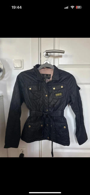 Svart quiltad jacka från Barbour - Quiltad svart jacka från Barbour med guldfärgade knappar, snörning i midjan och flera fickor. Klassisk look med Barbour-logga på bröstet och långärmad design. Perfekt för dig som gillar stilren och funktionell ytterjacka.