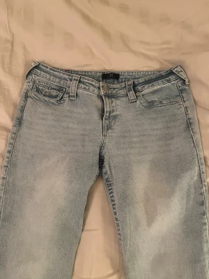 True Religion Joey Bootcut Jeans Ljusblå - Ljusblå jeans från True Religion, modell Joey Low Rise. Klassisk femficksdesign, låg midja och bootcut-ben. Jeansen är tillverkade i denim med snygg tvätt och har detaljerade sömmar samt dragkedja och knapp framtill.