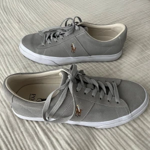 Grå mocka sneakers från Polo Ralph Lauren - Snygga grå sneakers i mocka från Polo Ralph Lauren med klassisk snörning och vit platt sula. Ikonisk broderad logga med ryttare på sidan och Polo-märke bak på hälen. Skorna har rund tå och mjuk innersida i beige ton. Mycket bra skick, använda max 2 gånger. Ordinariepris 1295kr