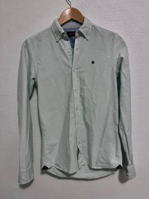 Mint grön skjorta från Morris XS - Fräsch mint grön skjorta från Morris i storlek XS. Klassisk modell med button-down krage, lång ärm och snygg broderad logga på bröstet. Skjortan är tillverkad i 100% mjuk bomull. Perfekt för dig som gillar stilrena och enkla plagg.