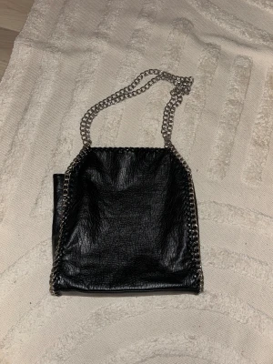 Svart axelväska från Tiamo med kedja - Cool svart axelväska från Tiamo i skinnimitation med grov silverkedja som axelrem och snygga detaljer längs kanterna. Väskan har ett innerfack med dragkedja. Sparsamt använd och i bra skick!
