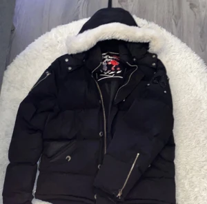 Moose knuckles  - Svart pufferjacka från Moose Knuckles med huva och vit fuskpäls runt kanten. Jackan har dragkedja, knappar och snygga detaljer på axeln. Perfekt för kalla dagar och har en cool streetstil.