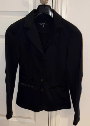 Svart kavaj från QHP med knappar - Snygg svart kavaj från QHP med klassisk krage och fyra knappar framtill och under så finns även en dragkedja. Jackan har två fickor med diskreta glittriga detaljer och är långärmad. Perfekt för dig som gillar stilrena och eleganta plagg med en liten twist. Den har inga skador eller repor.