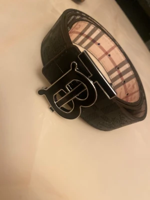 Burberry svart skärp med TB-spänne - Snyggt svart bälte från Burberry med klassiskt rutigt mönster på insidan och ett stort, glänsande TB-spänne i metall. Bältet är gjort i skinn och har en bred design som sticker ut. Perfekt för att lyfta vilken outfit som helst.