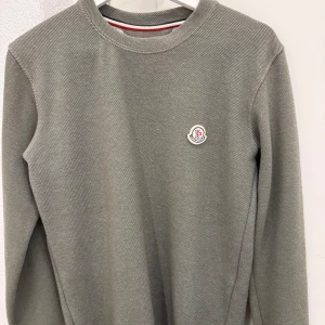 Grå långärmad tröja från Moncler - Snygg grå långärmad tröja från Moncler med diskret logotyp på bröstet. Tröjan har en rund hals och är tillverkad i mjukt bomullsmaterial. Perfekt för dig som gillar stilrena och klassiska plagg med exklusiv känsla.