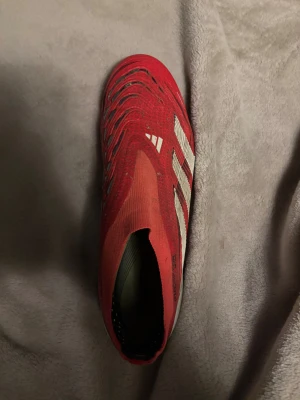 Adidas Predator röda fotbollsskor - Säljer ett par Adidas Predator fotbollsskor i rött och vitt med klassiska ränder på sidan. Skorna har en strumpliknande ovandel utan snörning och en platt sula med fasta dobbar för gräsplan. Materialet är syntet och designen är riktigt sportig och modern. Storlek 42 kommer också tvätta skorna innan don skickas för att få dom i lite bättre skick