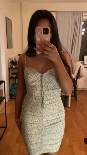 Blommig kort klänning med snörning - Säljer en söt kort klänning med blommigt mönster i ljusa toner. Klänningen har smala axelband, snörning framtill och en figurnära passform. Använts en gång. Säljs i befintlig skick. 