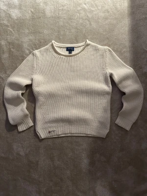Vit stickad tröja från Polo Ralph Lauren - Snygg vit stickad tröja från Polo Ralph Lauren med ribbad struktur och rund hals. Tröjan har långärmade muddar och en liten Polo-logga broderad vid nederkanten. Perfekt för dig som gillar klassisk och clean stil.