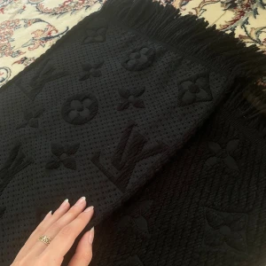 Svart Louis Vuitton halsduk i ull och silke - Exklusiv svart halsduk från Louis Vuitton med ikoniskt monogrammönster. Materialet är en lyxig mix av 94% ull och 6% silke, vilket gör den både mjuk och varm. Halsduken har snygga fransar i kanterna och en diskret, mörk look som passar perfekt till din vinterstil.