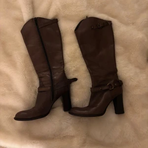 Bruna höga stövlar i skinn, storlek 37 - Snygga bruna stövlar i äkta skinn med hög klack och dekorativa spännen. Dragkedja på insidan och rund tå. Tillverkade i Italien, klassisk modell som passar till många outfits. 