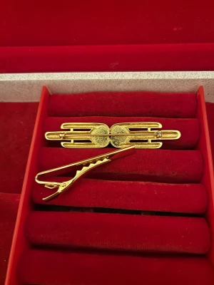 Guldpläterade slipsklämmor med hjärtan - Två stilrena slipsklämmor i guldpläterad metall. Den ena har två hjärtan dekorerade med små glittriga stenar och silverdetaljer, medan den andra har en minimalistisk design med guld och silver. Perfekt för att ge din outfit en extra touch.