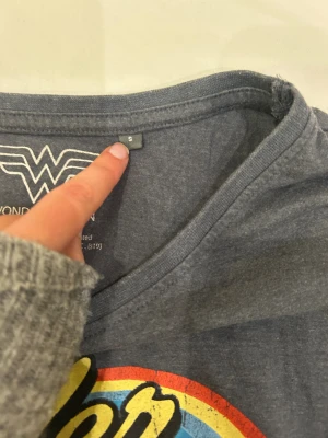 Wonder Woman t-shirt med regnbågstryck - Mörkgrå t-shirt med färgglatt Wonder Woman-tryck i gult, blått, rött och vitt på bröstet. Rund halsringning och korta ärmar. Mjuk och skön i bomullsmaterial, perfekt för dig som gillar superhjältar och retrostil.