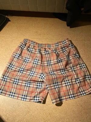Burberry rutiga shorts i bomull - Snygga Burberry shorts med klassiskt rutig mönster i beige, blått, vitt och rött. Modellen har elastisk midja och är tillverkad i mjuk bomull med lite stretch för extra komfort. Perfekt för dig som vill ha en clean och ikonisk look i sommar.