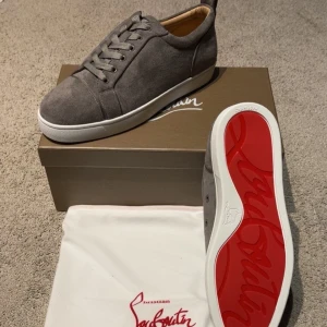Grå vita Louboutin skor - Snygga grå sneakers från Christian Louboutin i mjuk mocka med klassisk röd sula. Skorna har rund tå, snörning och vit platt sula. Perfekta för dig som vill ha en stilren och lyxig look med en touch av streetstyle.