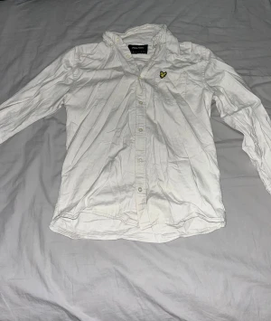 Vit skjorta från Lyle & Scott, storlek S - Vit skjorta från Lyle & Scott med klassisk krage och knappar hela vägen. På bröstet sitter den ikoniska gula fågeln broderad. Skjortan har lång ärm och är gjord i mjuk bomull, perfekt för en clean och stilren look. Pris kan diskuteras 