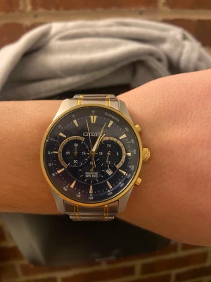 Citizen herrklocka i silver och guld - Citizen herrklocka med blå urtavla, kronograf och datumvisning. Armbandet är i rostfritt stål med guldfärgade detaljer och lås med Citizen-logga. Klockan har rund form, guldtonad boett och tre subdials för extra funktioner. Perfekt för dig som gillar stilrena accessoarer.