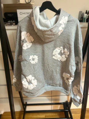 Grå hoodie med vita blommor Denim Tears - Grå hoodie från Denim Tears med vita blommiga tryck över hela tröjan. Tröjan har huva, magficka och ribbade muddar vid ärmslut och nederkant. Oversized passform och mjukt material som känns skönt mot huden. Perfekt för dig som gillar streetwear och unik design.