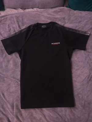 Svart McKenzie t-shirt i polyester - Svart t-shirt från McKenzie med diskret logga på bröstet och tonade detaljer längs axlarna. Tillverkad i mjuk polyester som ger en sportig känsla. Klassisk passform med rund hals och korta ärmar. Perfekt för en clean och enkel stil.