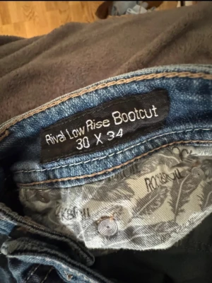 Rock & Roll bootcut jeans med broderi - Bootcut jeans från Rock & Roll som jag köpte second hand i London.i klassisk blå denim med låg midja. Framsidan har slitna detaljer vid knäna och baksidan har snygga broderade mönster på bakfickorna. Perfekt för dig som gillar en avslappnad och cool stil.