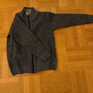 Verni-Grå stickad kofta med dragkedja - Mysig grå stickad kofta från Woolpower med hel dragkedja och ribbade muddar. Insidan är fodrad med mjuk fleece i rutig design med bruna, vita och grå toner. Perfekt för kyliga dagar och har två praktiska fickor med dragkedja. Den är aldrig använd vara provad säljer den för att det inte riktigt var min still. Den är lite mörkblå grå som färg 