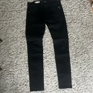 Svarta Replay Anbass jeans - Säljer ett par svarta Replay Anbass jeans i stretchigt Hyperflex-material. De har klassisk femficksdesign, smal passform och snygga detaljer vid fickorna. Perfekta för dig som gillar stilrena och bekväma jeans med modern look. Passar 170/175