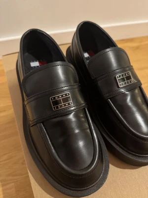 Nya svarta loafers Tommy Jeans skinn stl36 - Stilrena svarta loafers från Tommy Jeans i skinn med chunky sula och metallogo framtill. Klassisk design med rund tå och diskreta sömmar. Perfekta för dig som vill ha en clean och modern look med en touch av preppy vibe. Innersula mått: 22.9cm. Använda endast 1 gång, som nya! Köpte dem i november 2025. Nypris:1599kr