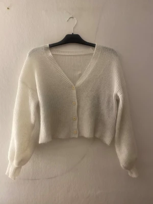 Vit croppad kofta med v-ringning - Säljer en vit, ribbad och croppad kofta med v-ringning och fyra knappar framtill. Koftan har långa ärmar och är stickad i ett mjukt material som passar perfekt för kyliga dagar. Enkel och stilren design som är lätt att matcha.