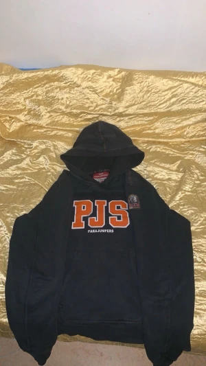 Svart hoodie från Parajumpers YL - Svart hoodie från Parajumpers med stor orange PJS-logga på bröstet och patch på axeln. Tröjan har huva med orange sömmar och är tillverkad i mjuk fleece. Klassisk känguruficka framtill och ribbade muddar. Perfekt för chill eller streetwear, har tappat lite färg men fungerar bra att använda.