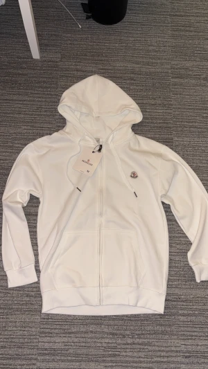 Vit hoodie från Moncler med dragkedja - Vit hoodie från Moncler med broderad logotyp på bröstet, huva med snörning och praktisk dragkedja framtill. Tröjan har två fickor och är tillverkad i mjuk bomull för skön komfort. Perfekt för dig som gillar stilrena och exklusiva plagg. Tveka inte för o skicka pris förslag